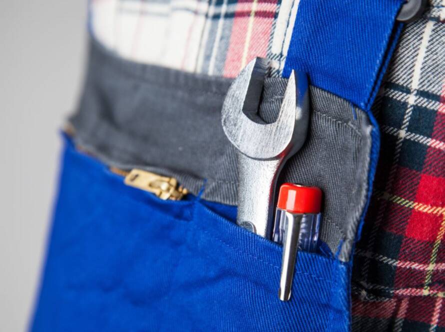 Das Bild zeigt eine Nahaufnahme eines Handwerkers in blauer Arbeitskleidung mit einem karierten Hemd. In der Werkzeugtasche befinden sich ein Schraubenschlüssel und ein Schraubendreher, die für verschiedene Reparatur- und Montagearbeiten verwendet werden.
