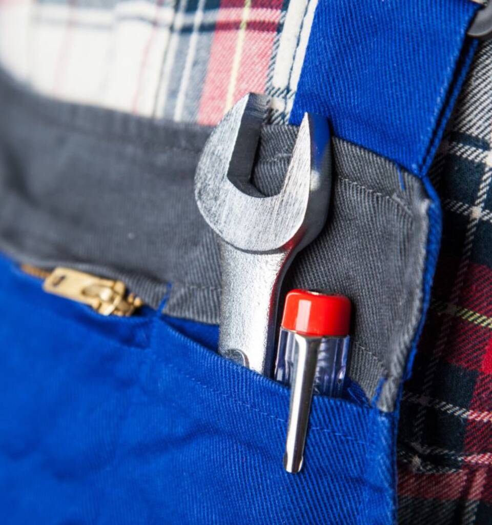 Das Bild zeigt eine Nahaufnahme eines Handwerkers in blauer Arbeitskleidung mit einem karierten Hemd. In der Werkzeugtasche befinden sich ein Schraubenschlüssel und ein Schraubendreher, die für verschiedene Reparatur- und Montagearbeiten verwendet werden.
